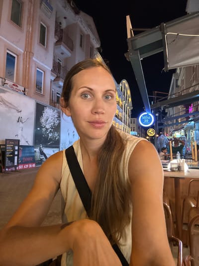 The media file is a portrait of a woman in an outdoor setting at night, possibly a street or promenade, given the illuminated signs and buildings. The woman is in the foreground, her face turned towards the camera, with a neutral expression. She has long, brown hair and is wearing a light-colored, sleeveless top with a dark strap across her shoulder.
Behind her, a building with multiple windows and balconies is visible, illuminated by warm light. Above the building, there are streetlights and neon signs, suggesting a commercial or entertainment area. One prominent blue neon sign reads "Most 10". Further in the background, there are hints of other people and structures, indicating a lively atmosphere.
The time of day is clearly night, characterized by the darkness of the sky and the artificial illumination. The weather appears to be clear. There are posters or large advertisements on the building, one of which depicts a tree. Visible text includes "Most 10" and a phone number "U 501 639 38 48" along with "DINER" and "Taba Çarşısı". The overall impression is of a casual evening outing in a Turkish town, possibly Fethiye.