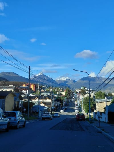 The image captures a daytime scene on a residential street in Ushuaia, Argentina, looking uphill towards a distant mountain range. The sky is largely clear blue with scattered white cumulus clouds. In the background, multiple mountains are visible, with several prominent peaks exhibiting snow cover.
The street itself is paved with asphalt, showing a darker, rectangular section in the mid-ground indicative of recent resurfacing or repair work. Vehicles are parked along both sides of the street. On the immediate left foreground, a light-colored minivan or car is partially visible, followed by a silver/light blue compact hatchback (consistent with a Citroën C3). Further up the street, a red SUV or crossover is parked on the right, near the resurfaced area, alongside other parked sedans and hatchbacks. No vehicles are in motion, and no occupants are visible.
Both sides of the street are lined with single- to two-story residential buildings of varying architectural styles and colors, including shades of yellow, beige, and brown. Overhead utility lines are prominent, strung between numerous wooden and metal utility poles, particularly on the left side of the frame. A streetlamp with a curved arm is positioned on the right side of the street in the mid-ground. Interspersed between the structures and along the roadside are various trees and shrubs. No individuals are depicted in the scene.