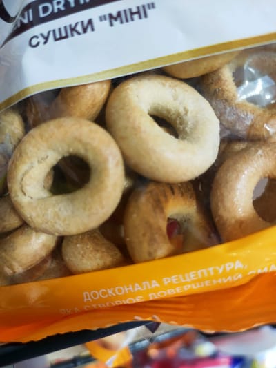 The image shows a close-up of a package of small, ring-shaped baked goods, likely "sushki" (small bagels or bread rings) based on the text visible on the packaging. The text on the white label at the top of the image is partially visible and appears to be in Cyrillic, reading "NI DRY" and "СУШКИ 'МІНІ'". The baked goods are golden brown and appear to be stacked inside a clear plastic wrapper. Below the baked goods, there is a bright orange band with more Cyrillic text, which is also partially visible and reads "ДОСКОНАЛА РЕЦЕПТУРА, СОВЕРШЕННИЙ СМАК". The image does not contain any information or visual cues that would allow for its localization to Kremenchuk, Ukraine.