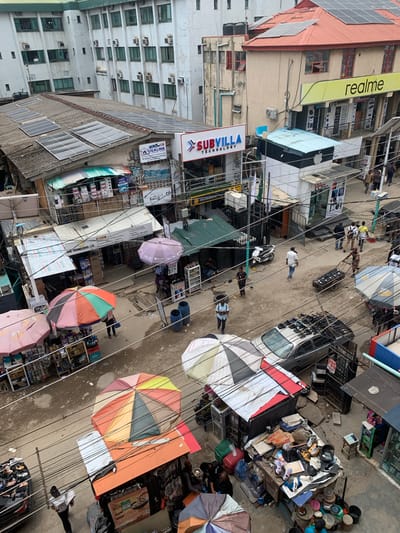 This is an aerial view of a street in Ikeja, Nigeria. The street is bustling with activity. People are walking and interacting with vendors under colorful umbrellas. The architecture consists of multi-story buildings with commercial signage, including brands like "realme" and "SUBVILLA TECHNOLOGY". Some buildings have solar panels installed on their roofs. Electrical wires crisscross above the street. Cars are parked along the street. One area includes what appears to be piles of used household items.