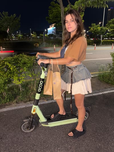 A young woman stands on a light green electric scooter at night in Incheon, South Korea. The scooter has the branding "GCOO" on its stem. The woman is wearing a cropped brown t-shirt, a denim skirt with a ruffled white hem, and black slides. She is holding two brown paper shopping bags. The background shows a street with lights, trees, and buildings. The scene appears to be in a city park or along a pedestrian walkway.
