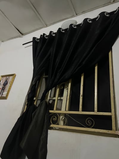 In a room in Howayi, Nigeria, black curtains with grommets are hung on a rod above a window. The window has a decorative gold metal grate with scrollwork. To the left of the window, a framed portrait of a man in a black suit is hanging on the wall. The ceiling appears to be made of white panels. The room is dimly lit, casting shadows on the walls and curtains.