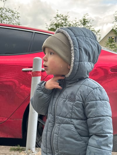 A young child, wearing a gray quilted jacket with the hood up and a gray beanie, stands near a red car. The child's right hand is near their face. A white and red striped pole is also visible. The scene appears to be outdoors, with trees and a building in the background. The location is identified as Akulovo (Gp Kubinka), Russia.