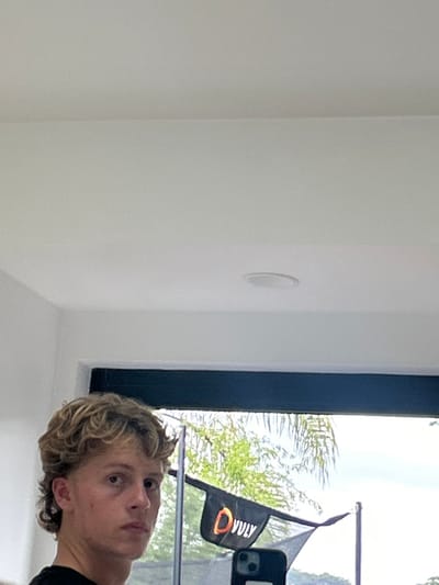 A person with curly blonde hair partially faces the camera, reflected in a window. The reflection also includes a black flag with the word "Vuly" on it. Outside the window, foliage is visible. The interior wall is white and features a circular vent or speaker.