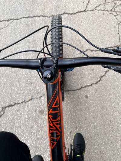 A first-person, downward-facing view captures the handlebars and upper frame of a black and orange Commencal "ALL MOUNTAIN" bicycle, situated on cracked light gray pavement in Cherven Bryag, Bulgaria. The bicycle features black handlebars, stem, and a top cap, with black control cables extending from the visible Shimano XT brake levers and shifters. Directly ahead, the knobby front tire is visible, indicating a mountain bike. The lower left of the frame shows a portion of a person's leg clad in black, while the lower right displays a person's right foot wearing a black sneaker with a neon green sole detail. The scene depicts a stationary bicycle with an individual standing near or over it on a paved surface.