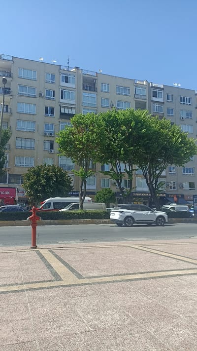The image depicts a street scene in the Yenişehir district of Turkey. A multi-story apartment building with a beige facade occupies the background. Several windows with white frames are visible, along with satellite dishes on the roof. In front of the building, lush green trees are in full foliage.
A street runs horizontally across the middle ground, with traffic visible. A white van with a red stripe is parked on the street, as is a white SUV. A bright red fire hydrant stands prominently on the sidewalk in the foreground, to the left of center. The sidewalk itself is paved with light-colored square tiles interspersed with darker and lighter colored decorative patterns. The sky above is clear and blue. The overall impression is of a sunny day in an urban environment.