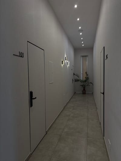 This media file captures a modern, empty hallway, likely within a residential or commercial building. The perspective looks down the length of the corridor, emphasizing its clean lines and contemporary design.
The dominant color palette consists of light greys and whites for the walls, floor, and ceiling, accented by black features. On the left wall, a series of small, circular recessed lights illuminate the space, complemented by three decorative wall fixtures further down. These fixtures are circular and appear to contain sparkling elements, possibly internal LED string lights, creating an inviting ambiance. Below these, a rectangular white access panel is visible. In the foreground on this wall is a light-colored door with a minimalist black handle, clearly marked with the black numbers "101".
The right wall is partially tiled with large, light grey square tiles from the floor up to a certain height, with a lighter painted wall above. Another rectangular panel, possibly an access hatch or vent, is visible near the top of this wall further down the corridor. The floor is also covered in matching light grey tiles.
At the far end of the hallway, a window with a light-colored roller blind provides a focal point. In front of the window stands a tall, potted palm-like plant, introducing a natural element to the otherwise stark environment. Adjacent to the plant is another door, similar in style to the one in the foreground, also featuring a black handle. The ceiling features black linear elements, possibly structural trim or part of a track lighting system.
The scene is devoid of people or activity, highlighting the architectural and interior design features. The artificial lighting suggests an indoor setting where time of day is less relevant. The overall impression is one of modern simplicity and functional elegance.