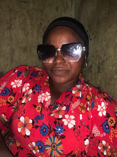 The image shows a person lying down, wearing a red dress with a floral pattern. The person is also wearing sunglasses and a black head covering. The background appears to be a textured, earthy surface, possibly a wall or floor. The lighting is dim, suggesting an indoor setting or a scene taken at night. The person's face is visible, but the sunglasses obscure their eyes. Their expression is neutral. The overall scene is still and lacks any overt action. There is no visible text in the image. The context provided indicates the location is Boi A, Nigeria.