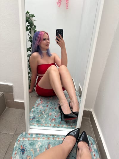A full-length mirror reflects a young woman with pink and purple hair. She is wearing a red outfit and black high heels. The mirror is in what appears to be a bathroom with tile flooring. A light blue rug with insect patterns lies on the floor. The reflection shows a white wall decorated with ivy and artificial flowers. There is a glimpse of what appears to be more tile or a threshold outside the mirror's frame. The location could be a private residence. The city is City of Zagreb, Croatia.