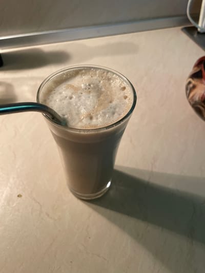 A tall glass of a foamy, light brown beverage, likely a milkshake or smoothie, sits on a light-colored, textured countertop. A metallic straw is partially submerged in the drink. The glass is positioned at an angle, with the top of the drink and the straw prominently featured. A dark shadow, cast by an unseen object, stretches across the countertop to the right of the glass. In the upper left corner, a sliver of what appears to be a stainless steel appliance is visible. In the background, a blurred object with red and black patterns is present. The overall lighting suggests it is daytime.