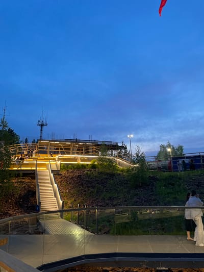 The image shows an outdoor amphitheater or viewing platform in Krasnoyarsk, Russia, at dusk. The structure is made of wood and metal, with illuminated stairs and railings. Several people are scattered throughout the area, some sitting on the steps and others standing and conversing. In the background, there is a construction site with scaffolding and a tall antenna tower. A red flag is visible in the top right corner of the image. The sky is a deep blue, indicating the transition from day to night, and the surrounding area is covered in greenery and rocks.