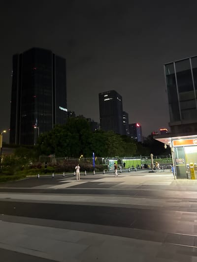 The image captures a nighttime scene in a modern urban environment, likely a plaza or walkway in Huangpu District, China. Dominating the background are several tall, dark skyscrapers with illuminated windows. In the foreground, a pedestrian walkway is marked by contrasting light and dark paving stones, creating a geometric pattern.
Several individuals are visible in the middle ground. One person, dressed in light-colored clothing, stands alone in the walkway, looking towards the camera. Further back, near a fenced-off green area, a small group of people are gathered, some seated at tables, and others standing. Bicycles are parked nearby, suggesting a public transportation or recreational area.
To the right of the scene, a section of a modern building with large glass windows is visible. Adjacent to it is a small kiosk-like structure with yellow signage and a display of products, possibly a convenience store or vendor stall. The overall atmosphere is one of urban night life, with the city lights and structures illuminated against the dark sky.