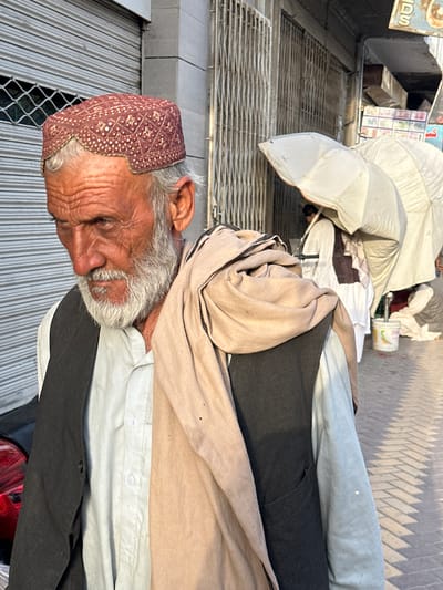 The image depicts an elderly man wearing a red and gold patterned skullcap. He has gray hair and beard, and his face is lined with age, giving him a thoughtful expression. He is dressed in traditional clothing, including a light-colored shirt and a dark vest. A tan-colored shawl is draped around his neck.

The man appears to be sitting or resting, possibly in an outdoor market or street setting. Behind him, there are indications of stalls or shops with metal shutters and a wire mesh fence. In the background, there are some scattered objects, including white fabric, possibly part of a stall or awning, and some boxes or crates. The overall scene suggests a casual daytime environment.

The lighting indicates it is daytime, with shadows cast on the ground, possibly indicating mid-morning or afternoon. The texture of the ground suggests it might be paved or a gritty surface. The man's posture and facial expression do not convey any strong emotions; he appears to be relaxed or perhaps contemplative.

There is some text visible in the upper left corner of the image on a sign, but it is partially obscured and difficult to read clearly. It appears to be in a script common in Pakistan. The broader context of Quetta City Tehsil, Pakistan, places the scene within a specific cultural and geographical setting.