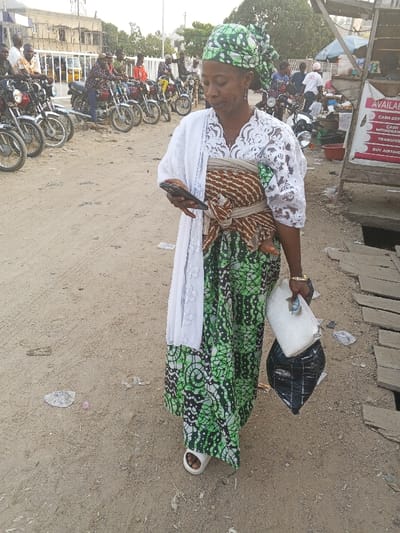 A woman stands in a dirt-covered area near motorbikes and a sign. She wears a green patterned skirt, a white lace blouse, and a green patterned headwrap. A brown and white patterned sling holds a baby on her front. She holds a smartphone in one hand and a white purse and a black plastic bag in the other. She wears white sandals. Buildings are visible in the background.