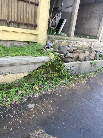 The image shows a section of a concrete sidewalk in Bandung, Indonesia, at the coordinates -6.938 latitude and 107.558 longitude. The sidewalk is cracked and uneven, with green plants growing up through the cracks. There are several rocks piled up next to the sidewalk. In the background, a yellow building with a barred window is visible. The sidewalk is adjacent to an asphalt road. A piece of yellow debris is visible in the lower left corner of the image. The concrete shows signs of weathering and erosion.