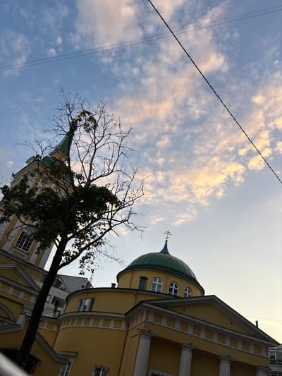 A low-angle, full shot captures the exterior of a yellow-colored church, possibly Orthodox, against a backdrop of a light blue sky with scattered clouds. The church features a green dome topped with a golden cross, arched windows, and white columns. A bare tree with a few green leaves branches across the left side of the frame, partially obscuring a section of the church's tall, spire-like structure, which also has a green roof. Overhead, power lines stretch diagonally across the sky. The architectural style and the presence of an Orthodox cross suggest the location might be in Eastern Europe. However, without further information or context from the image itself, definitively pinpointing the city as Riga, Latvia, is not possible solely from visual cues.