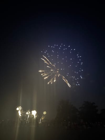 This is a night-time photograph of a fireworks display above a dark, hazy sky. In the upper right quadrant of the image, a large, fan-shaped burst of fireworks illuminates the sky with white and yellow sparks. Below this, towards the left and center of the image, a series of smaller, vertical bursts of yellow fireworks are visible, creating a cascading effect. A crowd of silhouetted people is gathered below, observing the display. Trees are visible in the lower right portion of the image, silhouetted against the hazy, dark sky. The fireworks are not located in the city of Lille, France.
