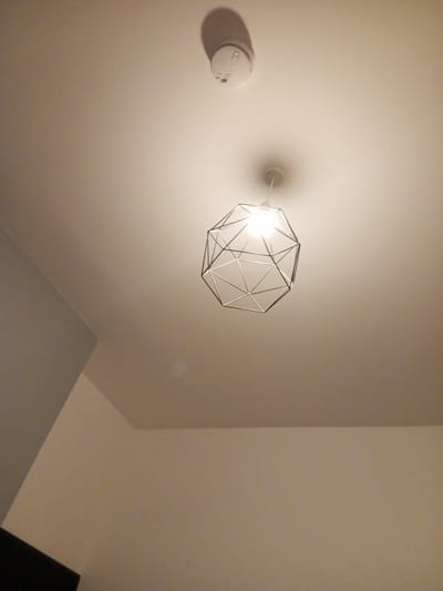 A wide-angle, low-angle shot captures a ceiling and upper portions of walls in a residential interior. The ceiling is painted white and appears smooth. In the upper left quadrant, a round, white smoke detector is mounted to the ceiling, casting a soft shadow. Below and to the right of the smoke detector, a modern pendant light fixture hangs from the ceiling. This fixture has a geometric, open-cage design constructed from thin, metallic rods, with a bright light bulb illuminating the interior of the cage. The walls are painted in neutral, light colors, with a visible corner where two walls meet in the upper left. The lighting suggests it is likely evening or indoors with artificial light sources. The scene does not provide any specific geographical markers to identify a location within Bristol, United Kingdom, or any other city. There are no people or animals present, and no actions or interactions are depicted. The environment is static and domestic.