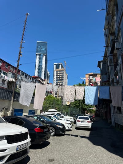 The image shows a narrow street in Batumi, Georgia, lined with parked cars, drying laundry strung between buildings, and modern high-rise buildings in the background. The street is paved and receives direct sunlight. Several cars are parked along the right side of the street, facing in the same direction. A Jeep with a French license plate is clearly visible. Laundry lines with hanging bedding are strung between buildings across the street. Tall, modern buildings are visible in the distance under a clear, blue sky. A construction crane is visible in the distance, possibly indicating ongoing construction. Buildings are visible on the left and appear to be older and more traditional in style compared to the high-rise buildings.