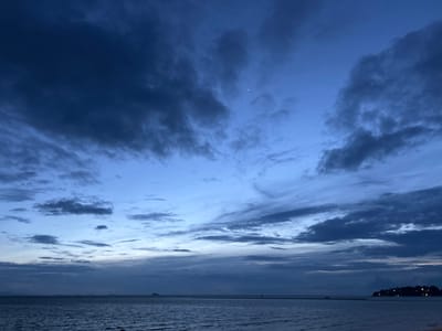 The image depicts a twilight scene over a calm body of water, likely the sea, near Ko Pha-ngan District, Thailand. The sky is a deep blue, transitioning to lighter shades near the horizon, and is filled with scattered clouds. Some clouds are dark and appear heavy, suggesting recent or impending rain, while others are wispy and illuminated by the fading sunlight. A sliver of the moon is visible in the upper right quadrant of the sky.
The water is dark and rippled, reflecting the dim light of the sky. The foreground shows a sandy beach. In the distance, a faint silhouette of land with lights can be seen on the right horizon, indicating a populated area. A narrow strip of land extends into the water from the right, creating a natural breakwater or sandbar. On this strip, a small, solitary figure is visible, appearing as a dark silhouette against the lighter background. Farther to the left, a small island or a distant landmass is barely visible on the horizon.
The overall mood is serene and tranquil, typical of the evening transition from day to night. The dim lighting suggests it is either early dawn or late dusk. The presence of the sea, beach, and the distant tropical landmass are strong cues for a coastal location in a warm climate. There is no visible text in the image.