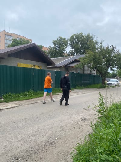 Two individuals are walking on a paved street in Poltava, Ukraine. The person on the left is wearing an orange t-shirt and blue shorts with athletic shoes. The person on the right is wearing a black hooded tracksuit and a black bag slung over their shoulder. They are walking away from the camera, towards the right side of the frame. The street appears to be in a residential area, with a green corrugated metal fence running along the left side of the road. Behind the fence, there are houses with dark roofs and some construction scaffolding. In the background, there are trees and a multi-story building. A white sedan is parked on the right side of the street, near a large tree. The sky is overcast.