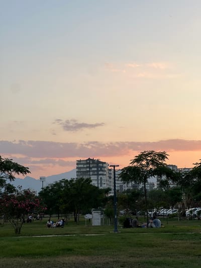 In a park in the city of Antalya, Turkey, a group of people are enjoying a pleasant evening. The sky is a soft gradient of blue, peach, and pink, with wispy clouds scattered across it. In the background, the silhouette of mountains can be seen, with a modern apartment building dominating the skyline in the middle distance. Several trees with lush green foliage are scattered throughout the park, providing shade and adding to the serene atmosphere. Some people are sitting on the grass, while others are lounging in chairs, engaged in conversation and enjoying the tranquil setting. The park also features a lamppost and a small white structure, possibly a playhouse or information booth. A parking lot with several cars is visible behind the trees, suggesting the park is accessible to the public.