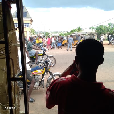The image shows a street scene in Nsugpe, Nigeria, viewed through a damaged window or doorway. Several people are gathered in the street, some of whom appear to be interacting or possibly arguing. Two motorcycles are visible in the foreground, one occupied. A three-wheeled vehicle (likely a "keke" or auto-rickshaw) is parked nearby. A building with solar panels is seen in the background. The individual in the foreground is possibly talking on the phone.