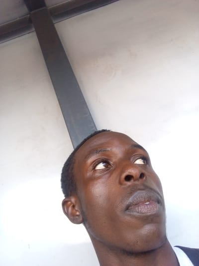 This is a close-up, low-angle, indoor photograph of a young Black male looking upwards. He has dark skin, short black hair, and is looking to his right and upwards. His eyes are wide open, and his lips are slightly parted. The background is a white wall with some faint staining and a dark, thick, horizontal beam that extends diagonally across the upper left portion of the image. The lighting suggests an indoor environment, possibly during the day. The overall mood is neutral. There is no visible text.