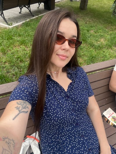 A young woman with long brown hair and sunglasses is sitting on a park bench in Pyatigorsk, Russia. She is wearing a dark blue dress with white polka dots. There is grass and trees in the background, suggesting an outdoor setting. She is holding her phone to take a selfie. A menu is on the bench next to her.