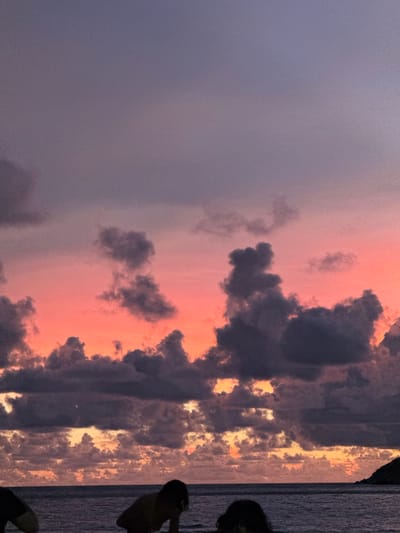 This is a photograph of a dramatic sunset over the ocean. The sky is filled with vibrant colors, ranging from deep purples and grays to warm oranges and pinks. Dark, heavy clouds are contrasted with brighter, illuminated patches of sky, creating a sense of depth and intensity. The sun appears to be setting on the right side of the frame, casting a golden glow on the clouds and the water.
On the right side of the image, silhouetted figures of people are visible, suggesting they are either standing on a beach or a pier, enjoying the sunset. The water stretches out towards the horizon, reflecting the colors of the sky. The overall scene evokes a sense of awe and tranquility, characteristic of a beautiful sunset.
The location is identified as Rawai, Thailand, which is known for its beautiful beaches and ocean views, making it a popular spot for sunset viewing. The time of day is clearly sunset, indicated by the warm, low-angled light and the colors in the sky. The weather appears to be clear enough for a sunset, despite the presence of dramatic cloud formations. There is no visible text in the image.