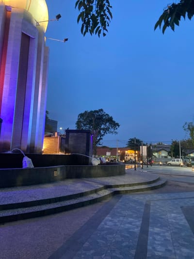The image shows a view of a fountain in the city of Sidoarjo, Indonesia, at coordinates -7.446 latitude and 112.719 longitude. The fountain is illuminated with purple and blue lights, and there are two jets of water shooting upwards from the base. The fountain is situated in a paved area, with steps leading down to a lower level. The surrounding area is dark, with only a few streetlights illuminating the scene. There are trees on the right side of the image, and a building in the background, illuminated with yellow light. In the distance, there are cars driving on a street with streetlights. The sky is a dark blue, with a few stars visible. The image was taken at dusk.