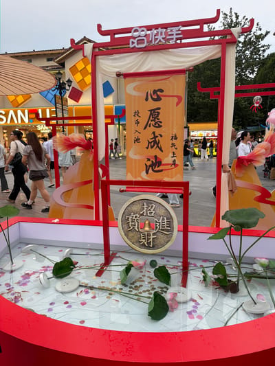 The image depicts a street scene in the Chongqing High-tech Zone, China, featuring a festive, Chinese-themed display. The central element is a vibrant red archway with ornate detailing, draped with sheer white fabric. Suspended from the arch is a large orange banner with black Chinese characters. Below the banner, a large, circular, antique-style coin with a bell and Chinese inscriptions is prominently displayed. The coin appears to be part of the decoration.

In the foreground, a shallow, red-rimmed pool is filled with water, decorated with lotus leaves, pink and purple paper cutouts, and small gold coins. Several tiny fish are visible swimming in the water, suggesting a "lucky fish" or similar attraction. White circular objects, possibly decorative elements or part of the display's mechanics, are submerged in the pool.

Behind the archway, a bustling street or marketplace is visible. People are walking around, some are looking at the display. Buildings with modern architecture, adorned with some decorative elements like checkered patterns, line the background. Signs in Chinese characters are visible on the buildings, including "SAN..." which is likely part of a store name. The overall atmosphere suggests an outdoor event or a decorated public space, possibly during a festival or celebration. The lighting suggests it is daytime, possibly late afternoon or early evening, with some shadows present.
