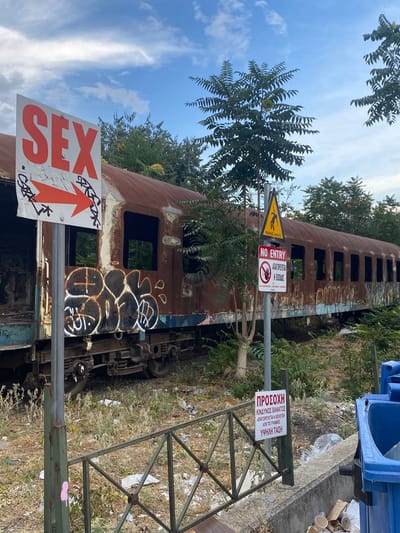 The photograph captures a scene with a derelict train car covered in rust and graffiti, situated in an outdoor environment. Several signs are present. A prominent white sign with red lettering reads "SEX" and features a red arrow pointing to the right. Another sign, circular with a red border, displays "NO ENTRY" in English and Greek text, along with a hand symbol. A yellow triangular sign with a black silhouette of a person falling is also visible. The train car is overgrown with vegetation, and the ground surrounding it is littered with debris, including plastic bottles and paper cups. In the background, trees and a cloudy sky are observable. The scene suggests an abandoned or disused area within an urban setting. There are no people in the frame.