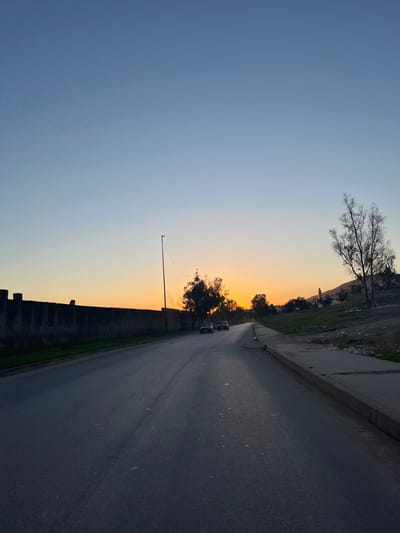 The image captures an outdoor scene at either sunrise or sunset, characterized by a dramatic gradient sky. When viewed correctly oriented (rotating the image 90 degrees counter-clockwise), the horizon line stretches across the mid-section, separating the sky from the land and road below.
The upper portion of the sky displays a clear, deep blue, gradually transitioning through lighter blues and soft purples to a vibrant orange and yellow hue closer to the horizon, indicative of either early morning or late evening. A bare, deciduous tree with intricate branches is silhouetted against the blue sky in the upper left quadrant.
Below the horizon, on the right side of the image, runs a dark asphalt road, appearing to be a two-lane path, leading into the distance. Two light-colored cars are visible on this road in the mid-ground, moving away from the viewer. Along the right edge of the road, a robust stone or concrete wall, dark and imposing against the lighter sky, runs parallel to the road, suggesting a boundary or a raised embankment.
To the left of the road and wall, several trees with denser foliage are silhouetted against the warm orange glow of the horizon. A tall, slender pole, possibly a utility or street light pole, stands upright in the mid-ground, rising from behind the wall. Beyond the trees and extending towards the upper left, there is a gently sloping landscape or hill, covered with sparse, dry vegetation. Faint outlines of what appear to be distant buildings or structures can be discerned on this hillside.
The overall mood of the image is serene and tranquil, with the warm colors of the sky creating a peaceful atmosphere. No discernable text is visible in the image.