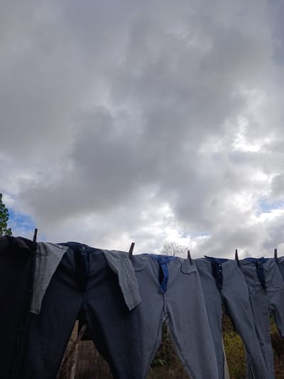 A line of jeans is hung on a clothesline outdoors, with a cloudy sky in the background. The jeans are mostly dark blue denim, with some lighter blue and grey fabric visible. They are attached to the line with clothespins. The environment appears to be rural or semi-rural, with some trees and vegetation visible below the clothesline. The image is taken from a low angle, looking up at the jeans and the sky. The location is specified as Altagracia, Venezuela.
