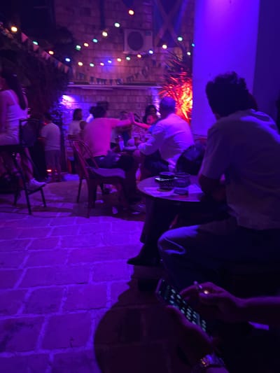 The image depicts an outdoor gathering at night, illuminated by colorful string lights and purple and red lighting, suggesting a bar or restaurant setting in Herceg Novi, Montenegro. Several individuals are present, some seated at tables, others standing. In the foreground, a person is holding a smartphone and what appears to be a cigarette. The ground is paved with stone tiles. The overall atmosphere is festive and social.