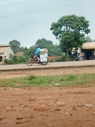 The image shows a man riding a motorcycle on a paved road in the town of Kivaywa, Kenya. The man is wearing a blue shirt and a hat, and the motorcycle is carrying a large load of packages. There is a second motorcycle with two riders in the distance, as well as a building and trees. The road is muddy and dusty. The image was taken from a point in the town of Kivaywa, Kenya,  at the coordinates latitude: 0.56, longitude: 34.806. The scene appears to be on the outskirts of town, with a single-story building visible behind the motorcycles. The man on the motorcycle is heading towards the viewer. The motorcycles are traveling at a speed that is consistent with travel on a town road. It is a quiet scene, and the only sound is the hum of the engines. The weather is overcast, but the air is still.