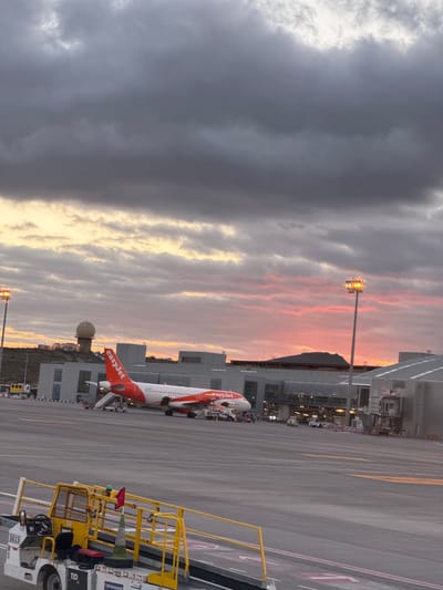 The image depicts an EasyJet airplane parked on the tarmac at Tenerife South Airport (TFS), located in Granadilla de Abona, Spain. A cloudy sky with hints of sunset colors forms the backdrop. A yellow service vehicle is positioned near the plane, indicating ground operations are in progress. The airport terminal building is visible in the background.