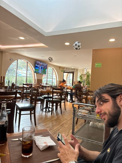 The image depicts the interior of a restaurant, likely a sports bar or themed eatery, possibly located in Cuautitlán Izcalli, Mexico, given the context. The decor features several soccer balls suspended from the ceiling. In the foreground, a young man with dark hair and a beard is looking at his smartphone, which he is holding in his hands. He is wearing a dark t-shirt with a golden emblem on it and a watch on his left wrist. A glass of dark beverage with ice and a bottle of Coca-Cola are on the table in front of him, along with a white napkin and cutlery. In the background, other patrons are seated at tables, and a large television screen displays a soccer match. The restaurant has a casual ambiance with wooden furniture and large arched windows offering a view of the outdoors.