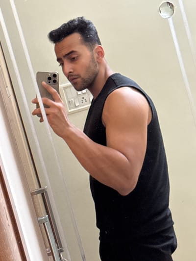 A man is taking a selfie in a mirror. He is wearing a black tank top and holding a smartphone. There is a light fixture on the ceiling. The reflection shows the man to be of South Asian descent. An electric outlet is visible on the wall behind him. The location cannot be determined as Kolkata, India from the image.