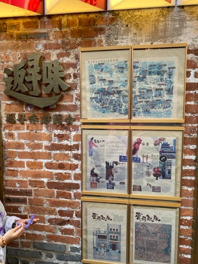 The media file captures an indoor scene dominated by a brick wall adorned with six framed artworks and decorative metal signs. The setting appears to be a cultural space or establishment in Guang Zhou Shi, China.

The upper portion of the brick wall features six rectangular frames arranged in two rows of three. The two frames on the left display illustrative, watercolor-style maps, depicting an intricate layout of buildings and streets. Visible text on these maps includes "您宁路永庆明名人目店地图" (You Ning Road Yong Qing Ming Celebrity Shop Map). The four frames on the right contain a collage of images, illustrations of traditional figures and objects, and extensive Chinese text, reminiscent of historical documents or old newspaper clippings. Phrases like "永庆" (Yong Qing), "YONGC," "大街" (Da Jie), and "ENG DAJE" are discernible, along with "YONG CING GATE" in one instance. Another piece of text, "咏春" (Yong Chun), is also visible, potentially related to one of the framed pieces or the metal signs.

Below the framed artwork, a large, intricately carved or cast metal character in a golden hue is mounted on the brick wall. To its right, a series of smaller, similar metal characters form a phrase. In the bottom right corner of the image, a person's hands are visible, holding a purple, braided object, possibly a decorative string or small toy. A beaded bracelet is also worn on one wrist. The overall impression is one of a richly decorated space, possibly highlighting local history, geography, or cultural heritage. The time of day and weather are not discernible.