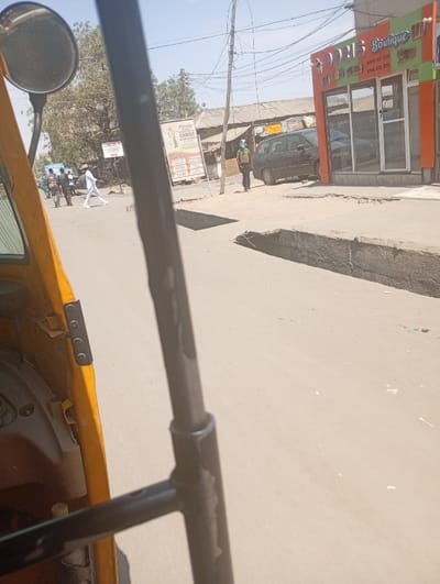 The image depicts a street scene in Sabon Gari East, Nigeria, viewed from the interior of a yellow tricycle (Keke Napep), partially obscured by a black metal structural bar in the foreground. The observer's round side mirror is visible in the upper left.

On the right side of the street, a two-story commercial building with an orange and white facade houses "DORIS Boutique," indicated by prominent signage including phone numbers. The building features large glass doors and windows reflecting the environment. A dark-colored minivan or small SUV is parked curb-side in front of this boutique. Standing on the pavement near the parked vehicle is a male individual wearing a light-colored helmet or headwear and a multi-colored vest, looking towards the left. The pavement on this side exhibits a partially damaged concrete drain or raised curb.

Across the street, on the left, several individuals are walking. One male figure in all white clothing (shirt and trousers) is prominent, crossing from left to right. Further down, multiple utility poles with a dense network of overhead electrical wires are visible. In the background, older, lower-rise buildings with corrugated metal roofs are present, along with large advertising billboards, one of which mentions "CHRIST." A large tree with sparse foliage stands on the left. The road surface appears to be asphalt or packed dirt, with a clear, sunny sky overhead.