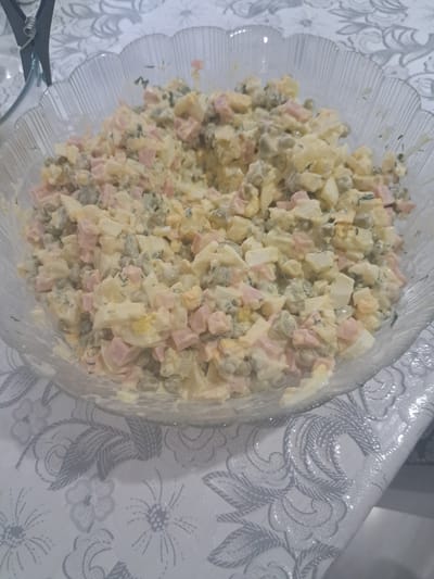 A clear, ribbed glass bowl is filled with a mixed salad. The salad appears to be composed of chopped ham, hard-boiled eggs, peas, and possibly potatoes, all coated in a creamy dressing. Fresh dill is also visible throughout the salad. The bowl sits on a white tablecloth with a grey, floral pattern. In the upper left corner of the image, a part of a grey metal object, possibly a binder clip, is visible. The scene is set indoors, with soft lighting. No specific location in Leningradskaya, Russia can be determined from the visual information provided.