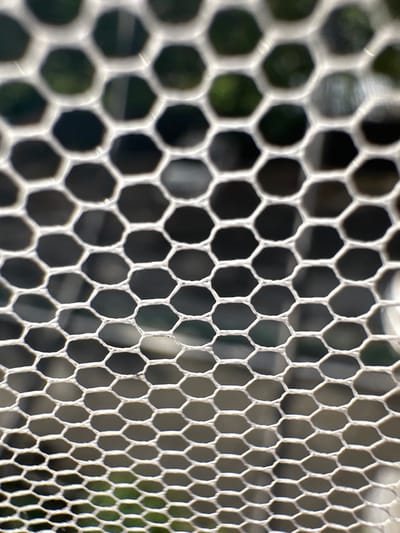 This is a close-up, abstract macro photograph of a hexagonal mesh or grid. The mesh is white and appears to be made of a woven material. The hexagonal openings are out of focus, revealing a dark, blurry background with hints of green, suggesting foliage or an outdoor setting. The perspective is angled, with the mesh receding into the distance and becoming more densely packed towards the right side of the frame. The lighting creates highlights on the edges of the mesh, giving it a slightly textured and dimensional appearance. There is no text visible in the image. The focus is entirely on the geometric pattern of the mesh and the abstract play of light and shadow through its openings. The scene does not depict any people, specific objects, or identifiable activities. The overall impression is one of texture, pattern, and depth created by the repeating hexagonal structure.