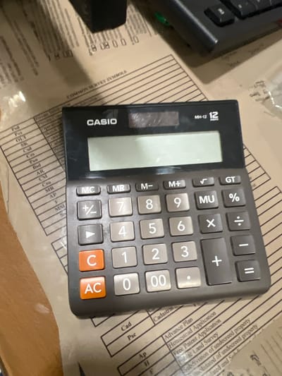 The image shows a close-up view of a Casio MH-12 calculator placed on top of a document. The document appears to be a printed form with columns and text, possibly related to land surveys or property records. The calculator has a light-colored display and a keypad with numerical and functional buttons. Some of the buttons visible include "MC", "MR", "M-", "M+", "√", "GT", "%", "7", "8", "9", "MU", "÷", "4", "5", "6", "X", "-", "1", "2", "3", "+", "=", "C", "AC", and "00".

There is no indication of the specific location within the city of Carmen, Philippines. The image does not contain any people or actions. The environment appears to be indoors, likely on a desk or table, with the lighting suggesting it was taken with a camera flash. The background shows more of the printed document and a dark object, possibly another electronic device like a keyboard.