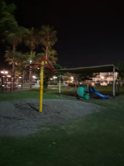 The image depicts a playground at night in an urban park setting in San Antonio, Peru. The playground features a merry-go-round with a yellow central pole and red circular frame, and a slide with green and blue elements situated under a shaded canopy. The ground is covered with sand in the immediate vicinity of the playground equipment, transitioning to grass further out. Several palm trees are visible throughout the scene, and streetlights illuminate the area, casting a dim glow. In the background, buildings with illuminated windows suggest a residential or commercial area adjacent to the park. The overall impression is one of a quiet, dimly lit public space.