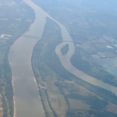 This aerial view shows a wide, meandering river, likely the Danube, flowing through an urban and semi-rural landscape. The riverbanks are lined with trees and some sandy areas. A long, narrow island with some vegetation is visible in the middle of the river. Several bridges cross the river, including a suspension bridge with tall towers and a simpler beam bridge further downriver. A white boat is sailing on the river. On either side of the river, there are areas of development, including buildings, roads, and agricultural fields. In the background, a hazy cityscape can be seen. The image is likely taken from a high altitude, possibly from an airplane. Based on the geography and bridges visible, this scene is consistent with the area near Dunakeszi, Hungary, where the Danube river flows and is crossed by multiple bridges.