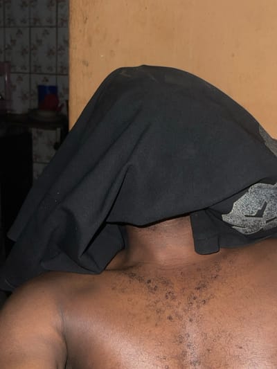 The image captures a close-up of a dark-skinned person's upper torso, partially covered by a black t-shirt with a lighter gray graphic on the shoulder. The bare chest area reveals scattered hair. The subject is in an indoor setting. In the lower left corner, there's a glimpse of a tiled wall with a floral pattern and some furniture or kitchenware beneath a counter.