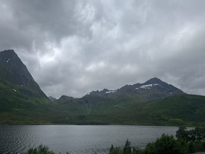 This is a horizontal, medium-shot photograph of a serene mountain lake in Gullesfjord, Norway. The sky is heavily overcast with thick, gray clouds. In the foreground, a dark, calm lake occupies the lower third of the frame. Its surface is rippled, reflecting the muted light. The middle ground is dominated by lush, green, tree-covered slopes that rise steeply from the water's edge. These slopes transition into rugged, dark-gray mountains in the background. Patches of snow are visible on the higher elevations of the mountains. The overall scene is one of natural grandeur and tranquility under a brooding sky. There are no people or animals visible in the frame. The image appears to have been taken from a slightly elevated vantage point on the shore of the lake.