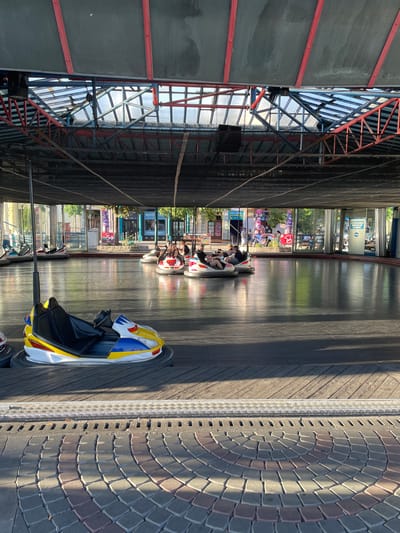 This image captures a scene at a funfair, likely in Vienna, Austria, with a focus on a bumper car ride. The ride is enclosed by a dark netting structure on the left, with exposed metal beams and translucent panels revealing a bright, sunny sky. On the right side, a textured pathway made of interlocking bricks forms a circular pattern, adjacent to a metallic strip and wooden planks.
Several bumper cars are visible on the slick, dark surface of the ride area. In the foreground, a yellow, blue, and white bumper car is parked, its black seat prominent. Further back, other bumper cars are in motion, carrying people. Some individuals are seated inside, seemingly enjoying the ride. The expressions of the people are not clearly discernible due to the distance and image quality.
Behind the bumper car track, a row of glass-enclosed booths or buildings lines the area, with trees and greenery visible beyond. Sunlight casts long shadows across the ride, indicating it is daytime, likely afternoon given the angle of the sun. The overall atmosphere appears to be one of lighthearted recreation.
There is some visible text on the buildings in the background, including what appears to be signage for "MAGIC" and other indecipherable words on posters. The bright and sunny weather contributes to a cheerful outdoor ambiance. The setting suggests a typical amusement park or fairground environment.